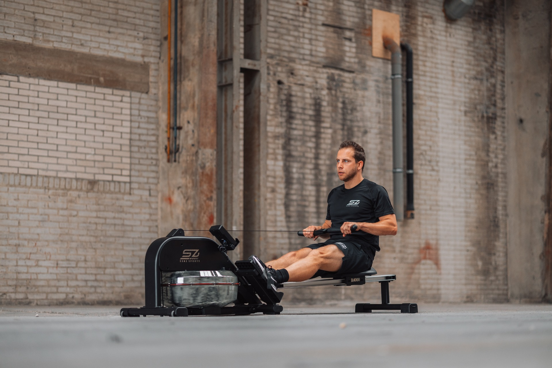 Man rowing on a machine indoors. He wears black clothing. The background features brick walls and industrial elements.