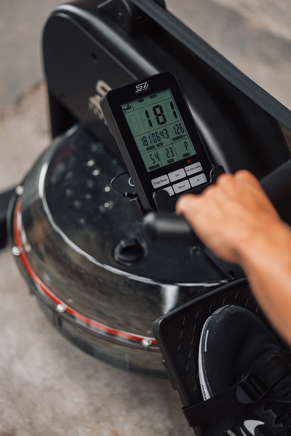 A person is using a rowing machine. The screen shows stats. A foot and hand are also visible.