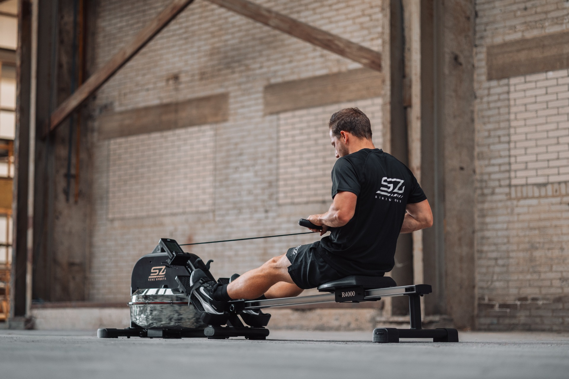 A man in black attire is rowing on a machine. The background is a brick wall and wooden beams.