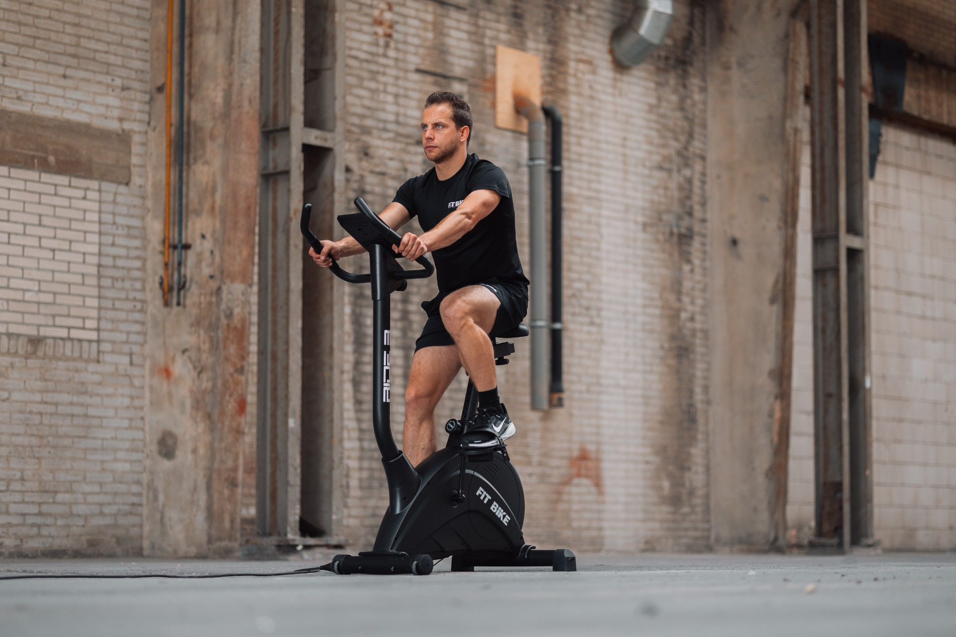 Homme en vêtements noirs sur un vélo d'appartement dans un cadre industriel. Murs en briques et en béton en arrière-plan.