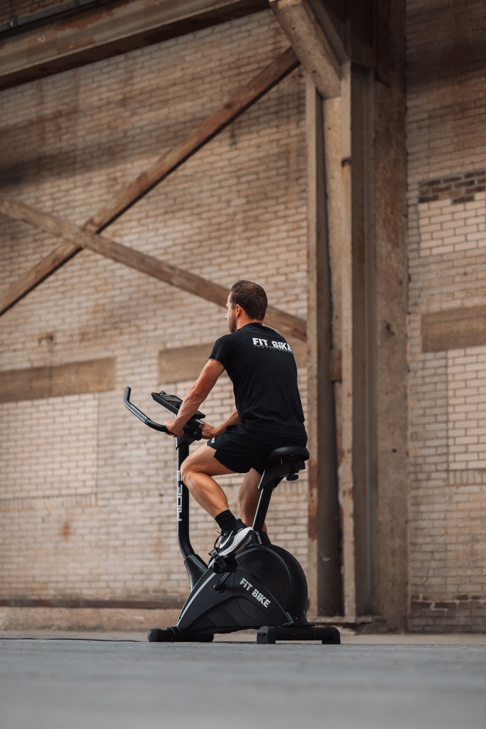 Un homme en vêtements noirs fait de l'exercice sur un vélo d'appartement à l'intérieur d'un bâtiment en briques. Le vélo dit 'FIT BIKE'.