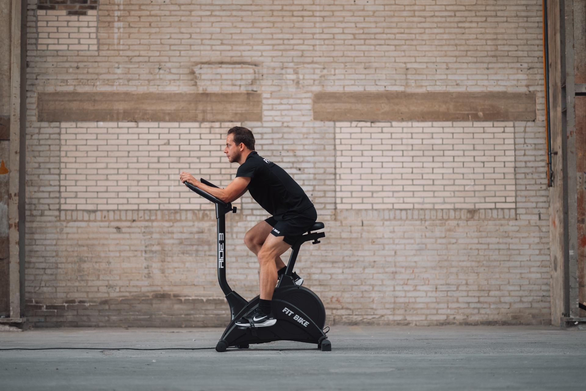 Homme sur un vélo d'appartement devant un mur de briques. Il porte un short et un t-shirt noirs. Le vélo indique FIT BIKE.