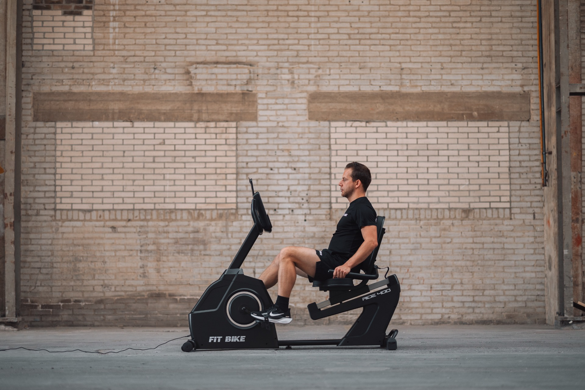 Un homme en vêtements noirs est sur un vélo d'appartement, contre un mur de briques. Le vélo est noir et porte la marque Fit Bike.