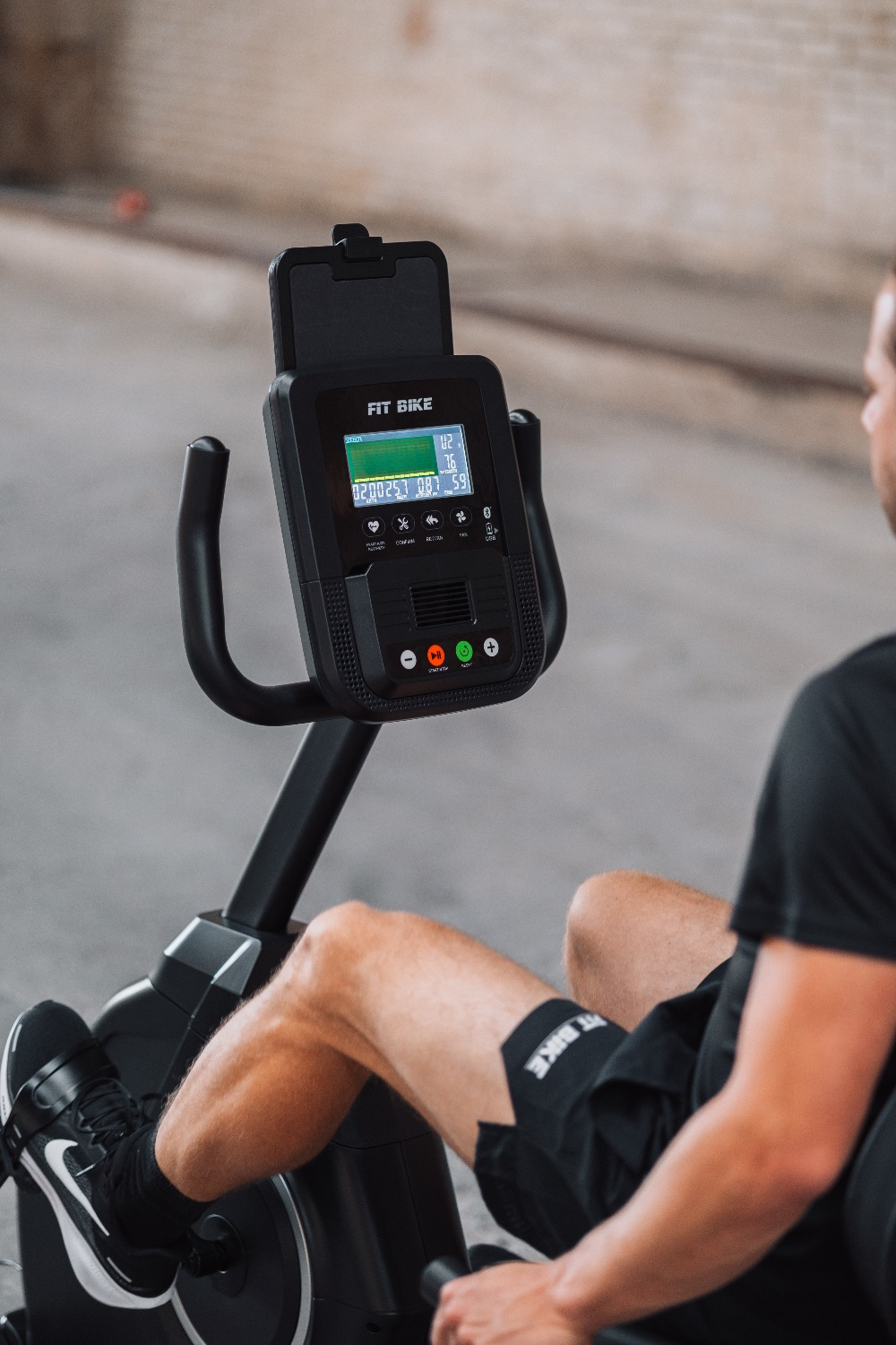 Un homme sur un Fit Bike. Vélo noir, écran affiche des données. Il porte un short et des chaussures noirs. Arrière-plan flou.
