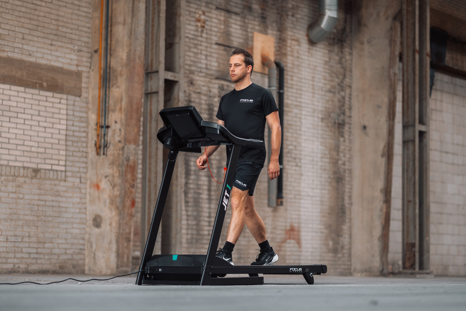 A man in black sportswear walks on a treadmill. The background shows a brick wall and industrial setting.