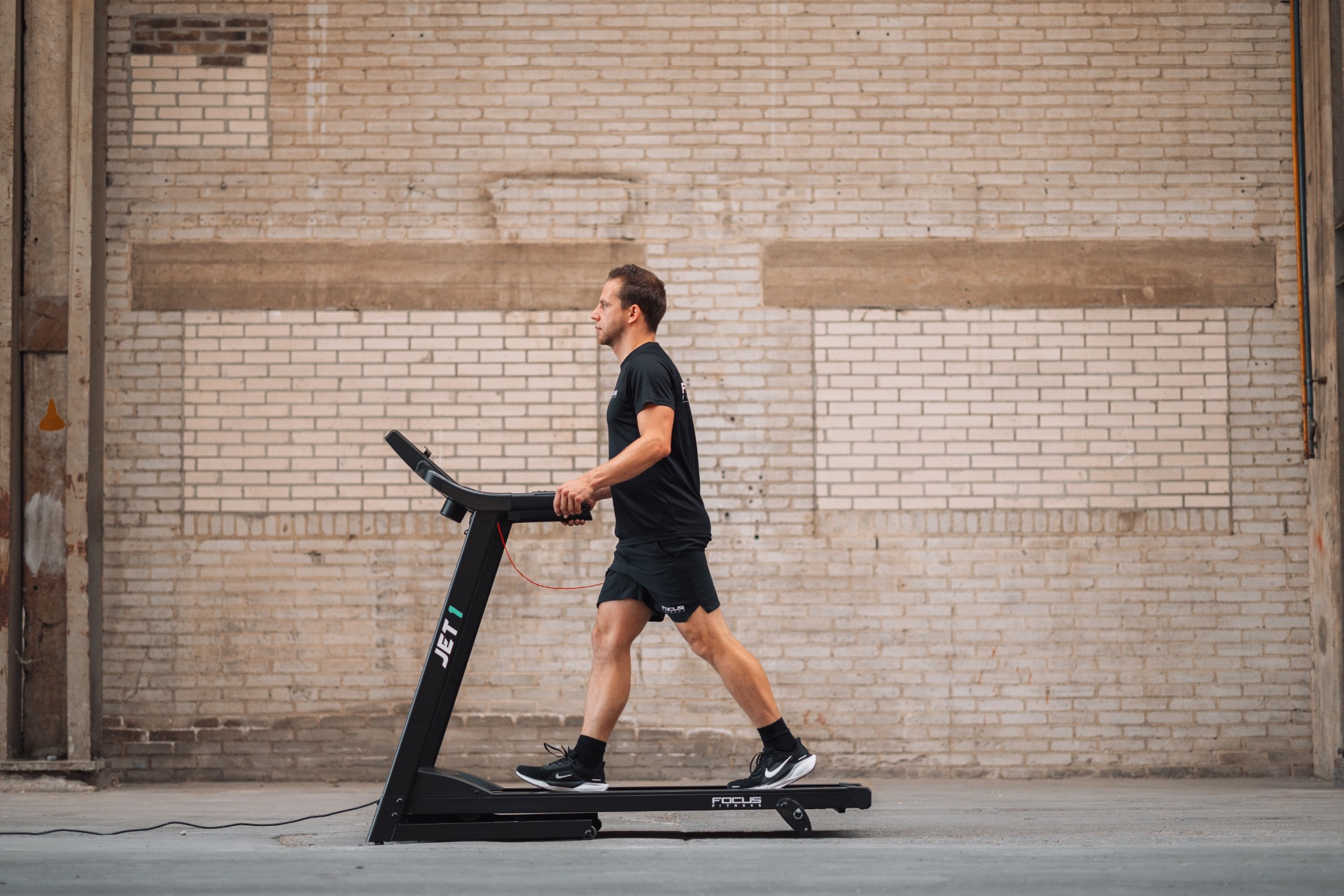 A man in black running on a treadmill in front of a brick wall.