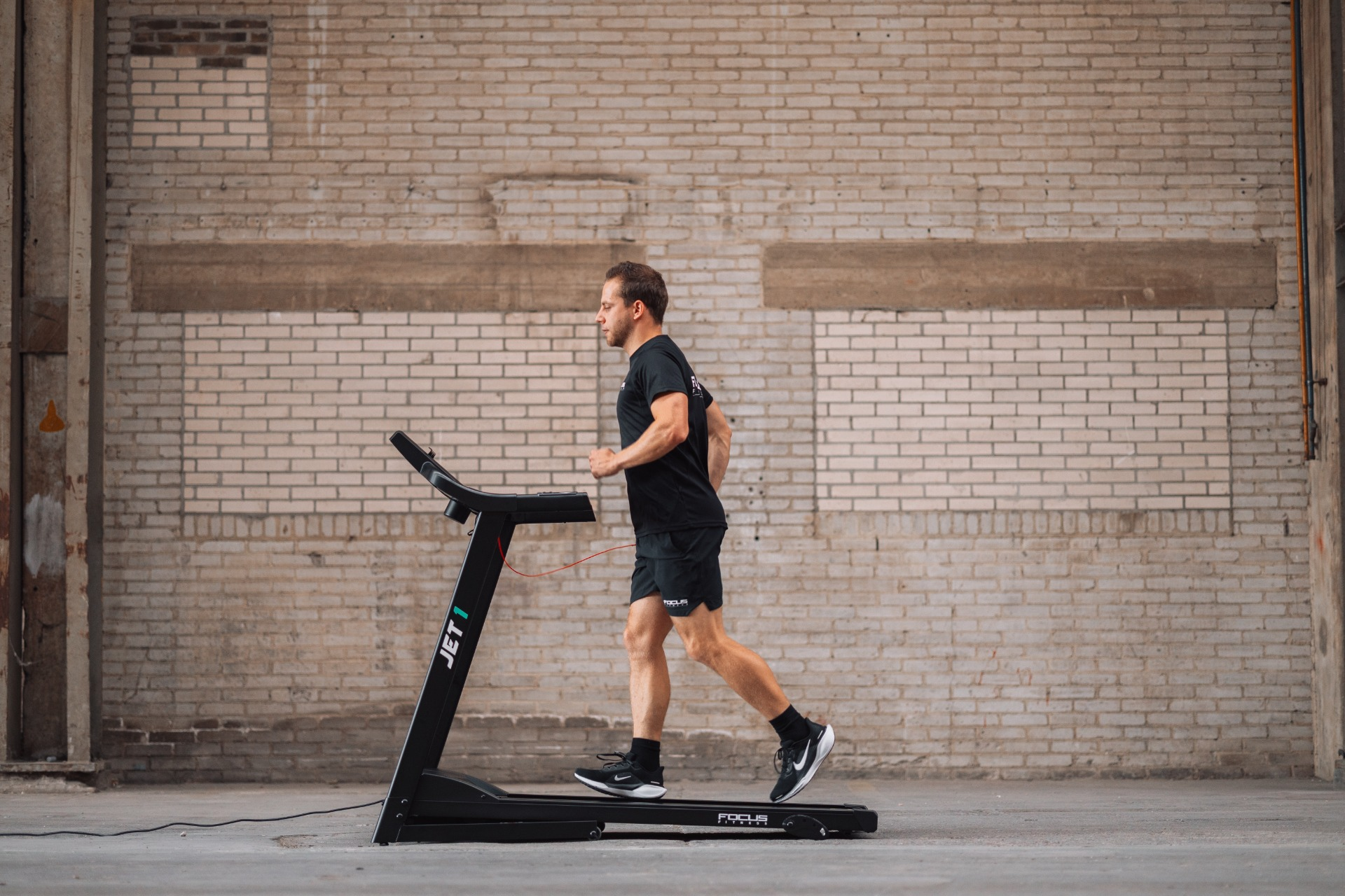 A man runs on a treadmill in a gym. He wears black shorts, t-shirt, and shoes. Brick wall in the background.