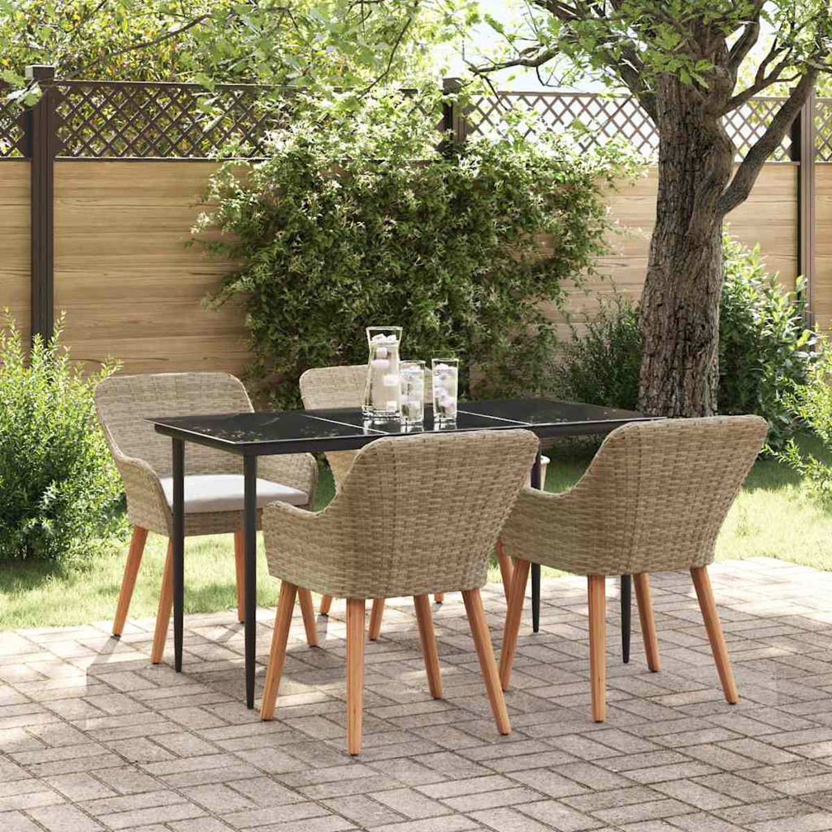 Essgruppe im Freien: schwarzer Tisch, Korbstühle, Gläser und Wasserkrug. Auf einer Ziegelterrasse mit Holzzaun und Grün.