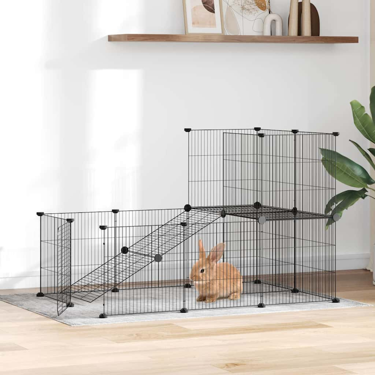 Ein braunes Kaninchen sitzt in einem schwarzen Drahtkäfig mit Rampe und Plattform. Der Käfig steht auf einer grauen Matte in einem Raum.