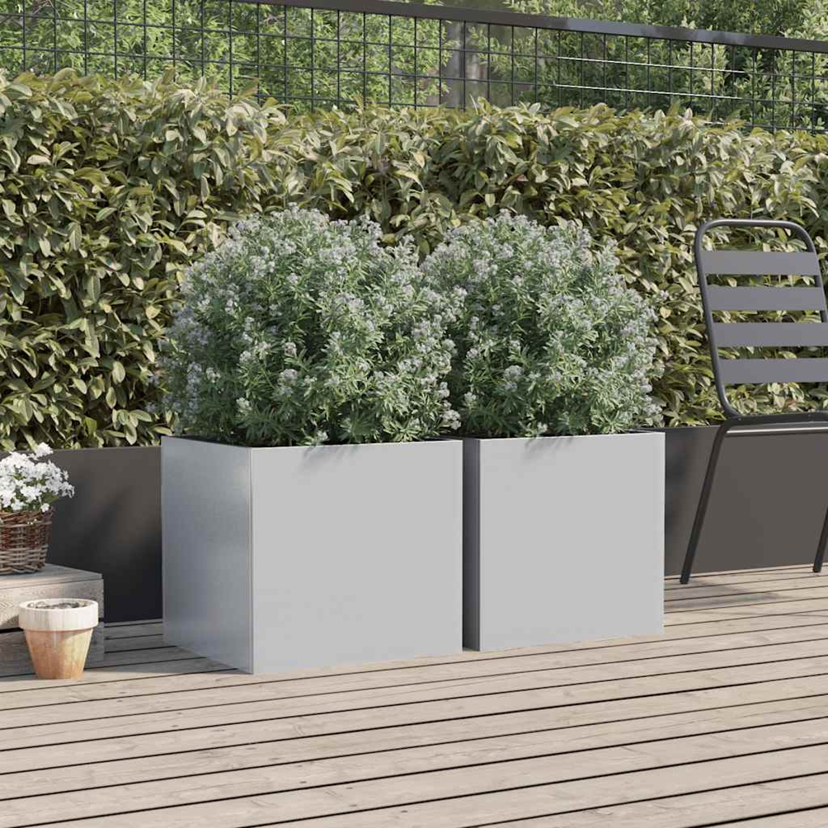 Zwei quadratische Pflanzgefäße mit grünen Pflanzen. Ein Stuhl und eine Hecke befinden sich im Hintergrund. Holzterrasse im Vordergrund.