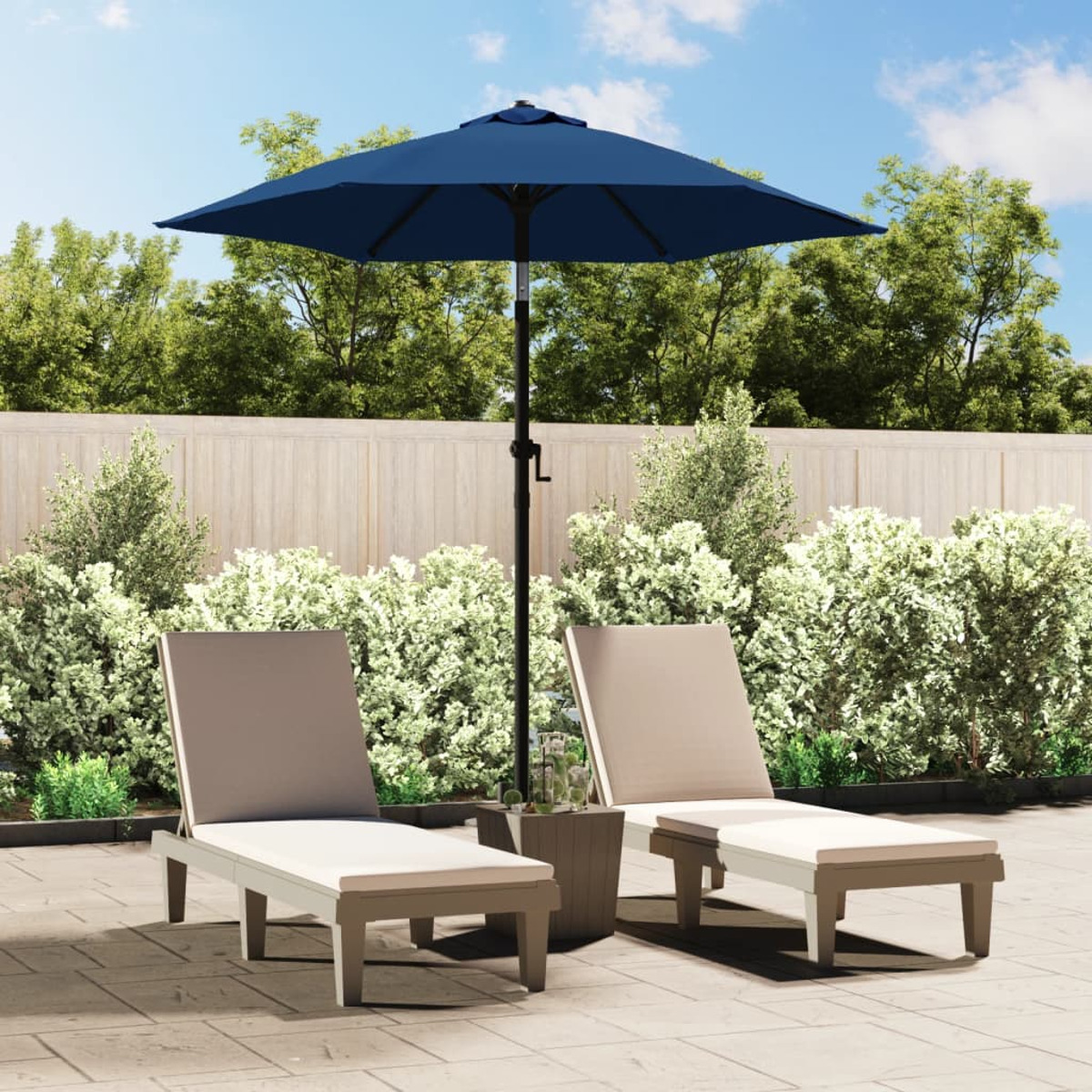 Due sedie a sdraio beige sotto un ombrello blu su un patio. Arbusti verdi e una staccionata in legno sullo sfondo.