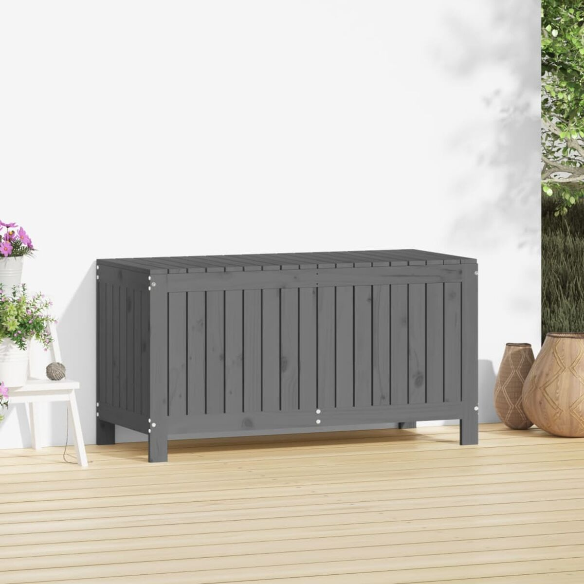Un baule di legno grigio su una terrazza di legno. Muro bianco sullo sfondo. Vasi e piante a destra e a sinistra.