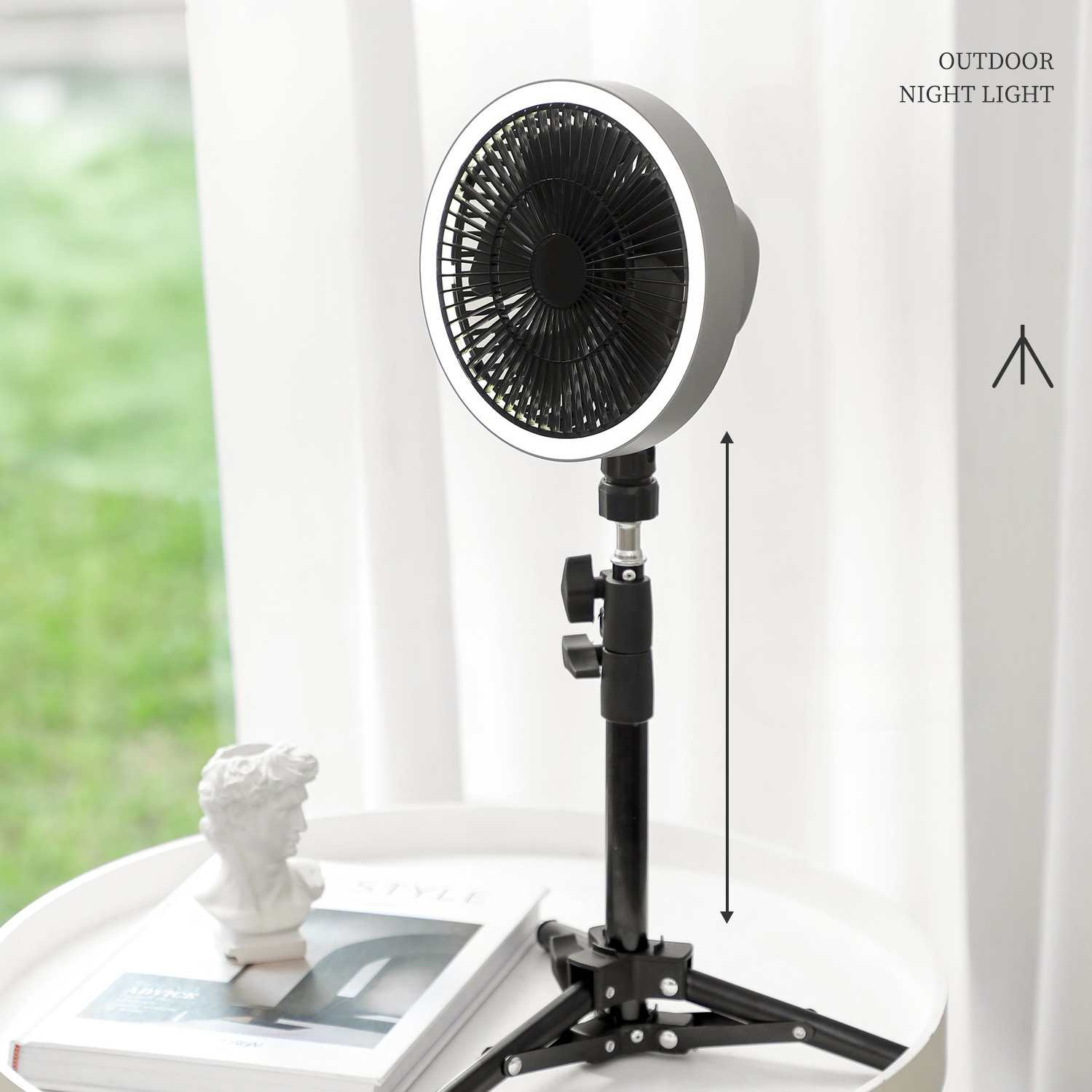 Ein kleiner Ventilator mit Lichtring auf einem Stativ, neben einer weißen Skulptur und einem Buch, in der Nähe eines Fensters.