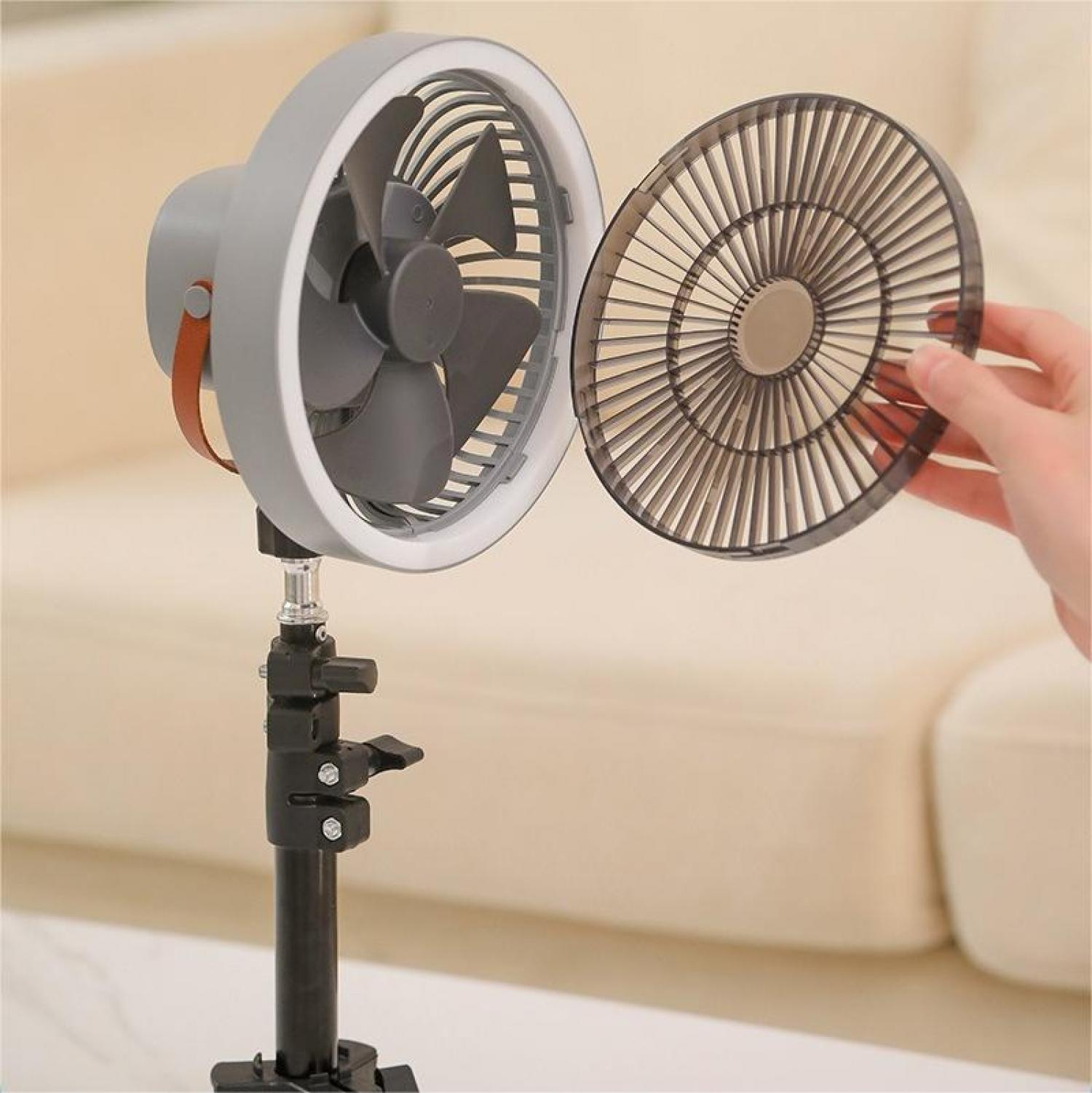 Ein grauer Ventilator mit abnehmbarer Abdeckung wird von einer Hand gehalten. Der Ventilator steht auf einem schwarzen Ständer. Hintergrund ist beige.