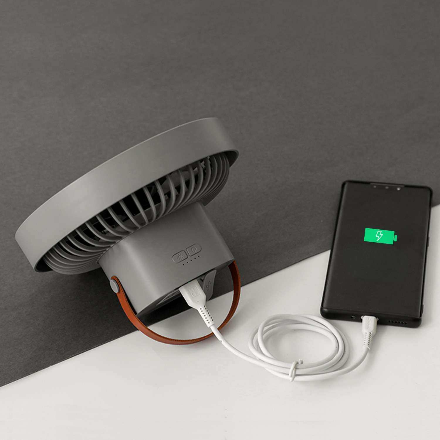 Ein grauer Ventilator mit braunem Griff, ein Telefon, das mit einem weißen Kabel geladen wird. Das Telefon zeigt ein Ladesymbol.