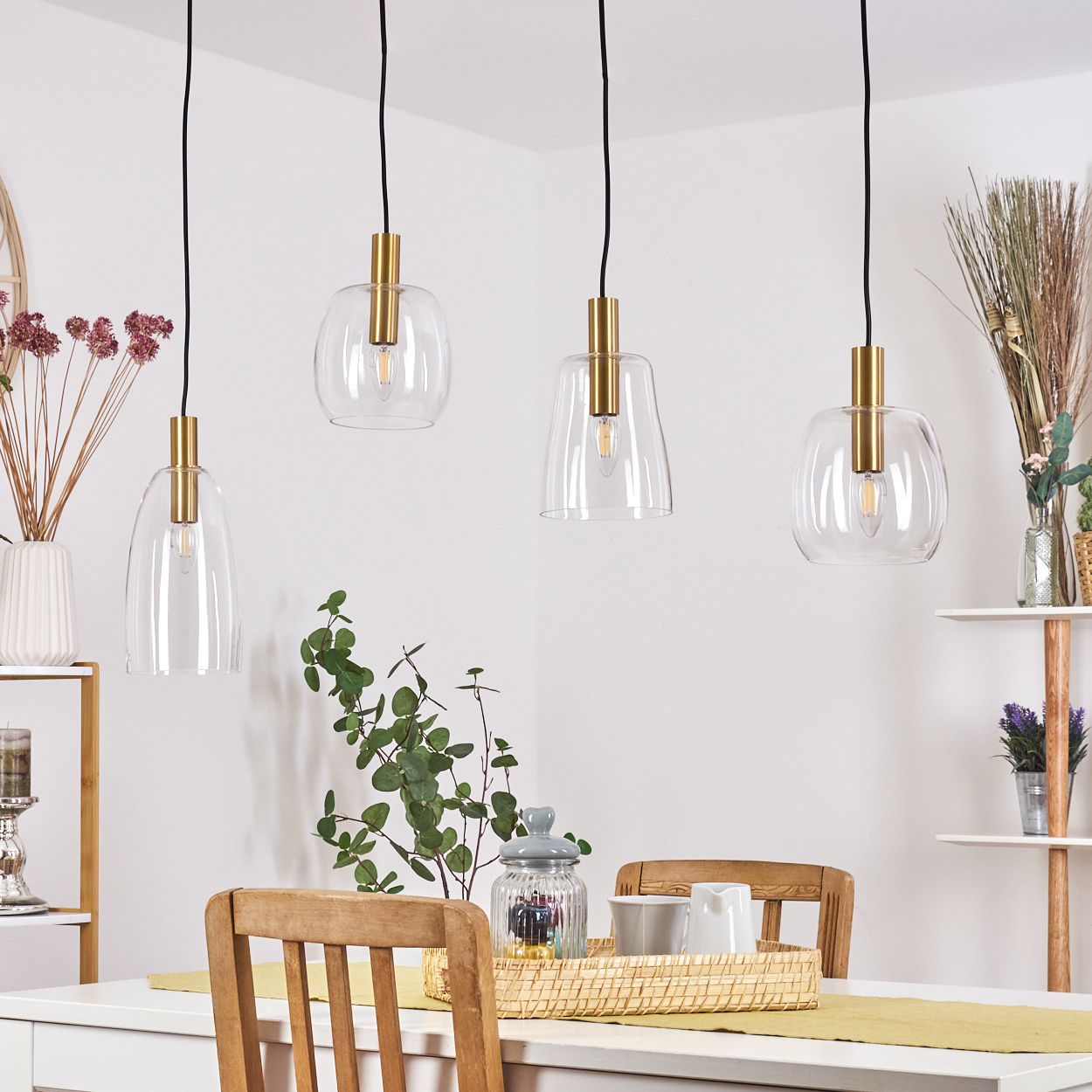 Vier Glas-Pendelleuchten hängen über einem Esstisch mit Holzstuhl und Dekorationsartikeln in einem hellen Raum.