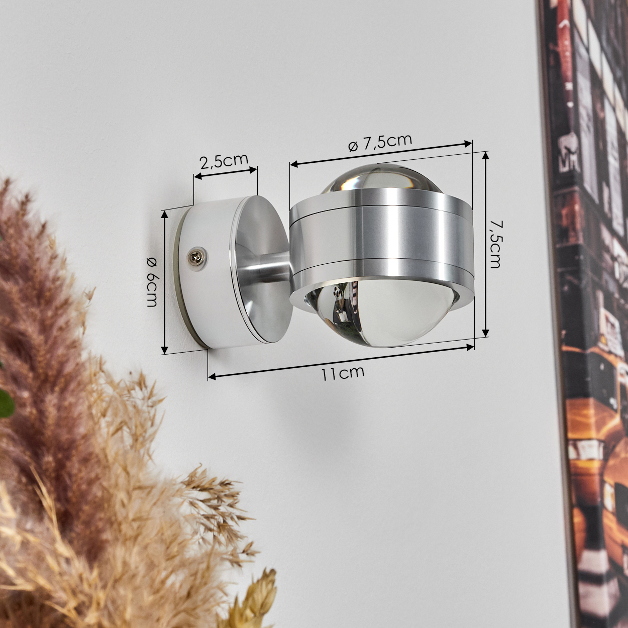 Moderne silberne Wandleuchte mit Maßangaben. Die Lampe ist an einer weißen Wand montiert, im Vordergrund dekorative Elemente.