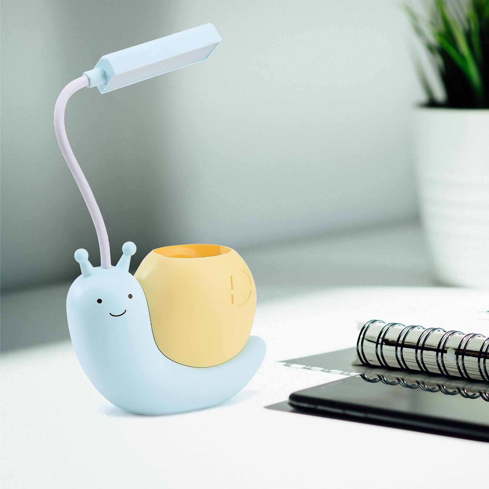Eine blaue, schneckenförmige Schreibtischlampe mit gelbem Gehäuse auf einem weißen Schreibtisch. Ein Notizbuch und eine Pflanze im Hintergrund.