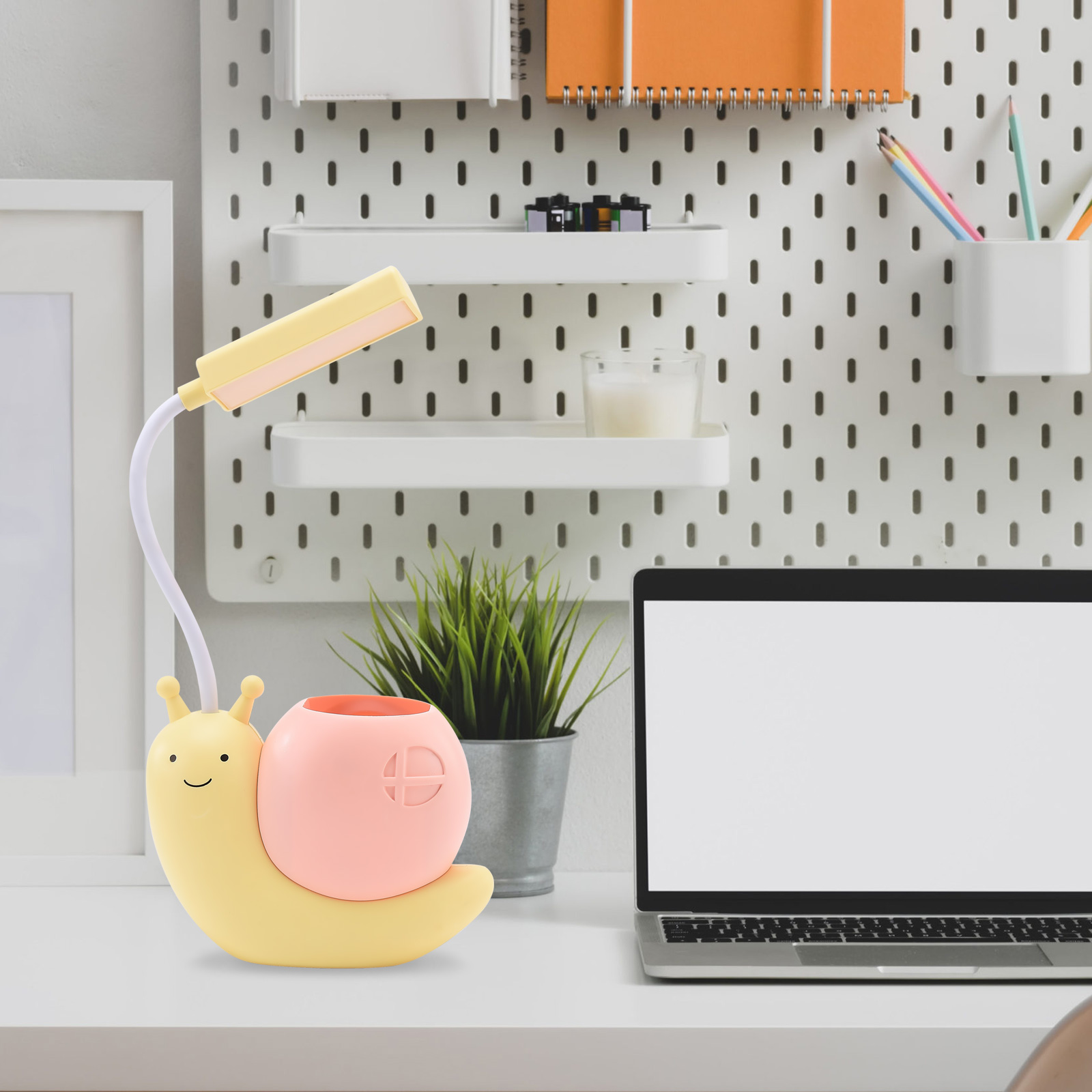 Ein Schreibtisch mit einer Schneckenlampe, einem Laptop und einer Pflanze. Die Lampe ist gelb und rosa. Ein Pegboard-Hintergrund ist sichtbar.
