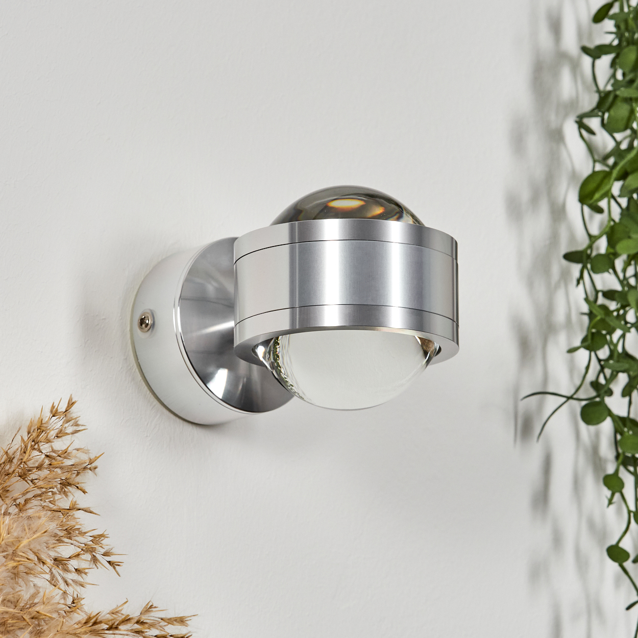 Eine moderne silberne Wandlampe mit einer kugelförmigen Glühbirne. Sie ist an einer weißen Wand befestigt, mit Blättern im Hintergrund und trockenem Gras im Vordergrund.