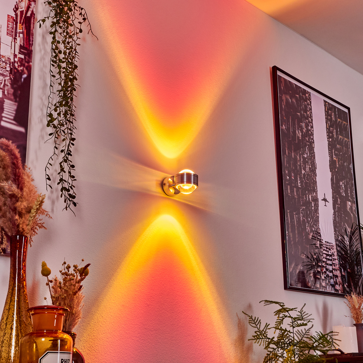 Eine Wandlampe wirft gelbes und rotes Licht. Ein Bild einer Stadt und Pflanzen sind sichtbar. Die Wand ist weiß.