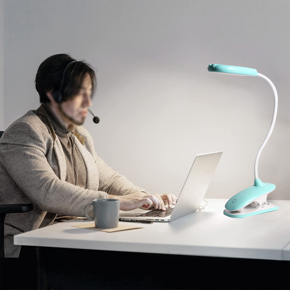 Person mit Headset tippt auf einem Laptop. Klemmleuchte davor. Tasse und Papier auf dem Schreibtisch. Hellblaue Lampe, weißer Schreibtisch, graue Wand.