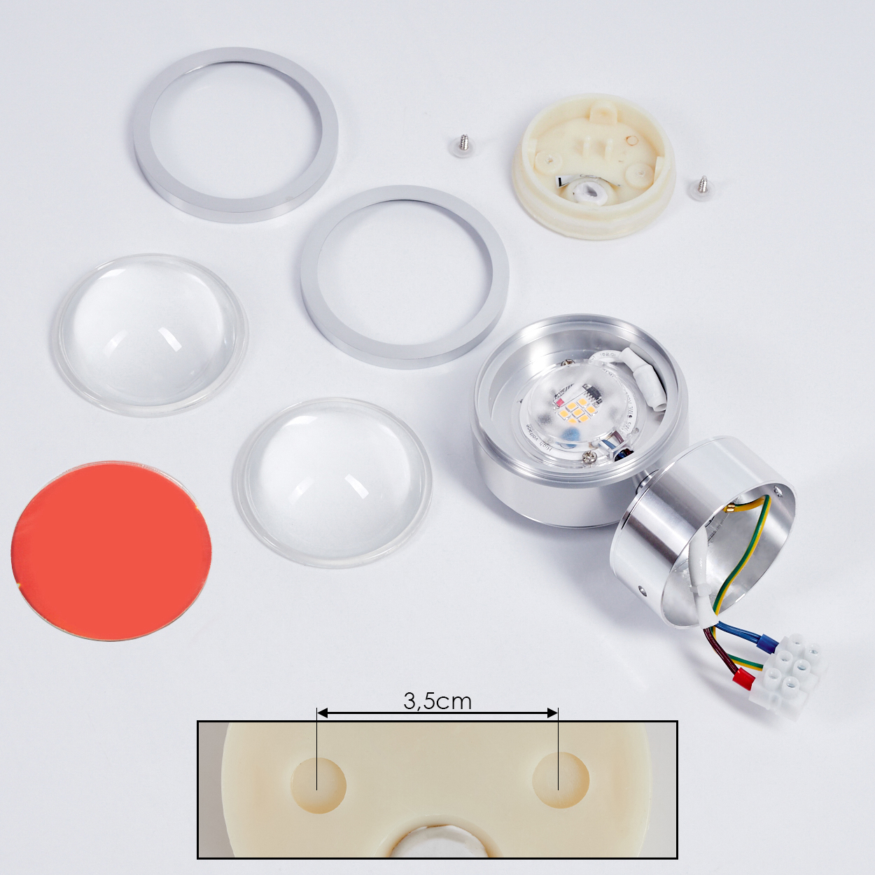 Zerlegte LED-Leuchtenkomponenten. Silberringe, klare Linsen, roter Kreis und Lichtbasis sind abgebildet. Drähte und eine Basis mit Löchern sind sichtbar.