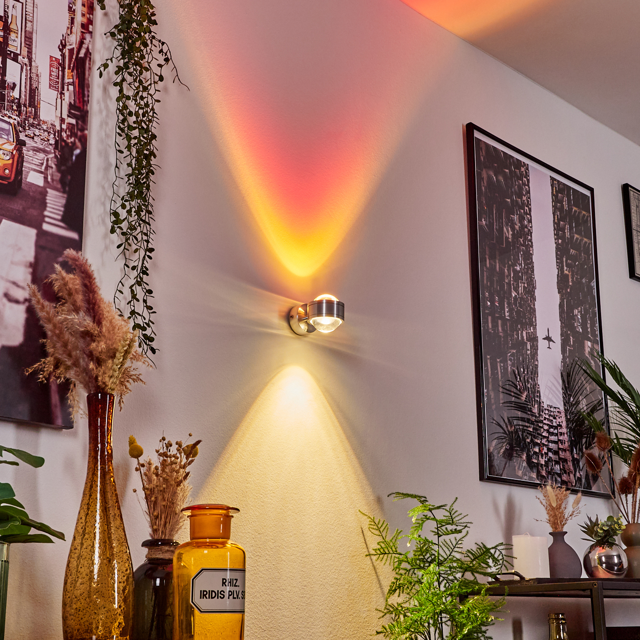 Eine Wand mit einer Leuchte, die orangefarbenes und gelbes Licht aussendet. Es gibt gerahmte Bilder und dekorative Vasen. Pflanzen im Vordergrund.