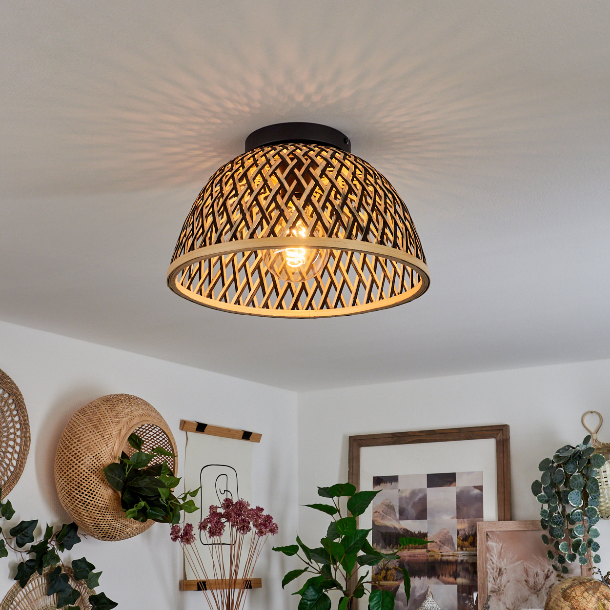 Eine gewebte Deckenlampe mit eingeschalteter Glühbirne hängt. Die Umgebung umfasst Pflanzen, Bilder und einen gewebten Wandkorb.