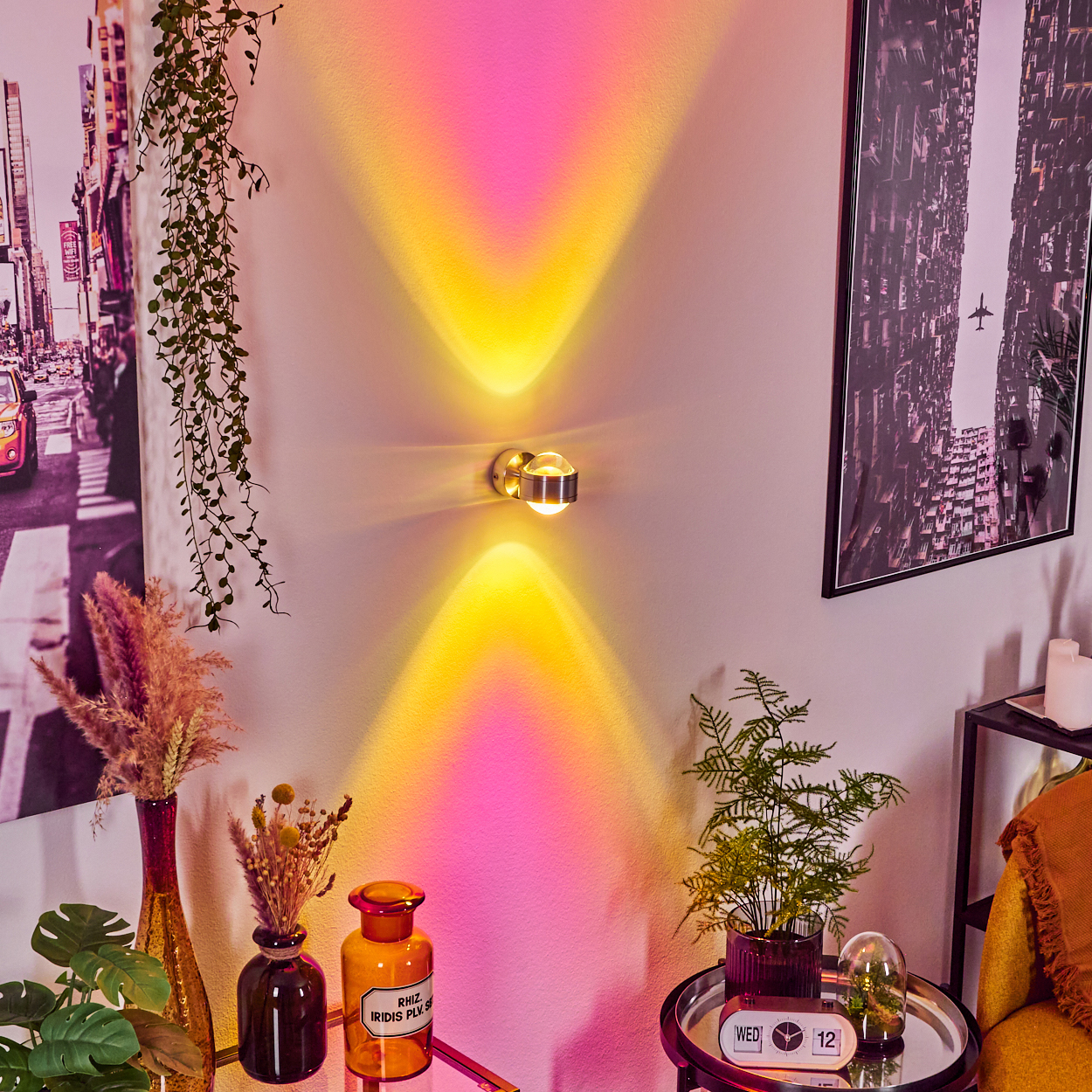Eine Wand mit einer Leuchte, die gelbes und rosa Licht wirft. Kunstwerke und Pflanzen schmücken die Wand. Ein kleiner Tisch trägt Pflanzen und eine Uhr.