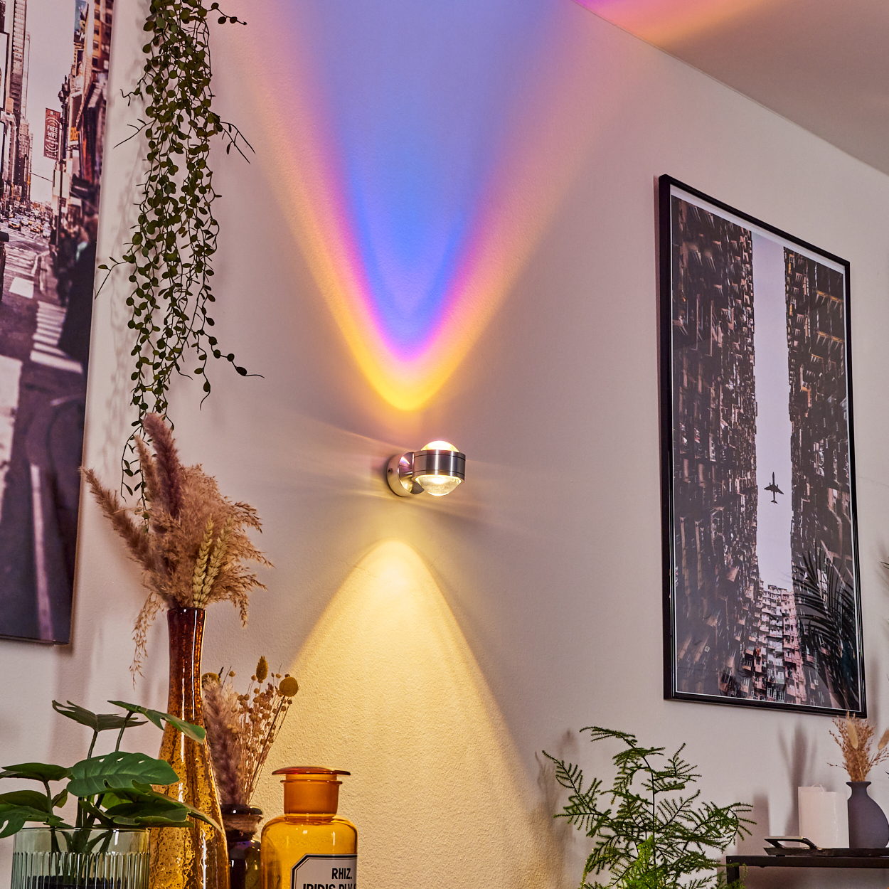 Eine Wand mit einer Lampe, die Regenbogenlicht projiziert. Im Vordergrund ein gerahmtes Bild, Pflanzen und Vasen.