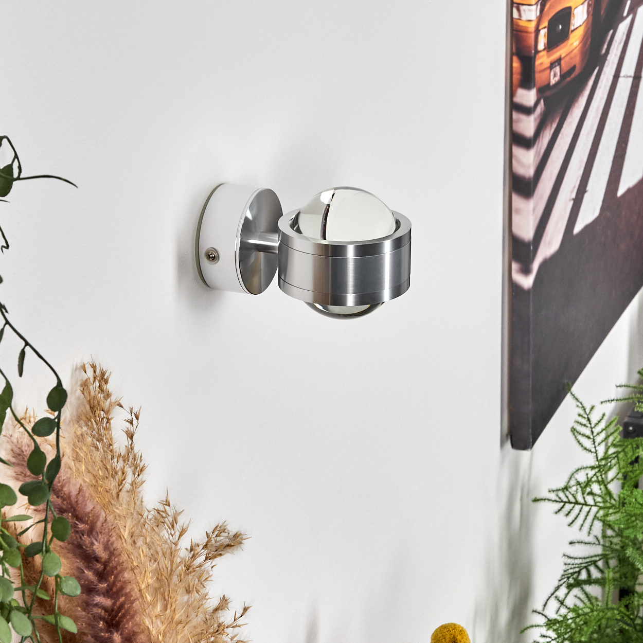 Eine moderne, metallische Wandlampe ist an einer weißen Wand befestigt. Rechts ist ein Bild eines gelben Busses. Getrocknete Pflanzen sind darunter.