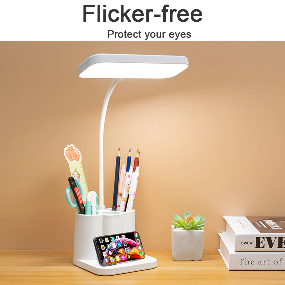 Eine weiße Schreibtischlampe mit Stifthalter, Bleistiften, einer Schere, einem Telefon, Büchern und einer kleinen Pflanze auf Holz.