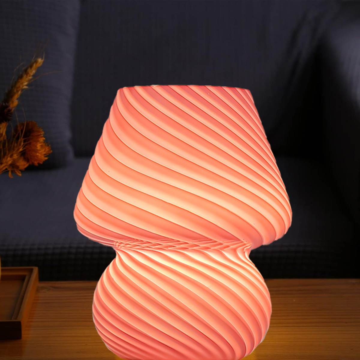 Eine pilzförmige Lampe mit Spiralmuster, die rosa leuchtet. Sie steht auf einer Holzoberfläche mit dunklem Hintergrund. Getrocknete Blumen links.