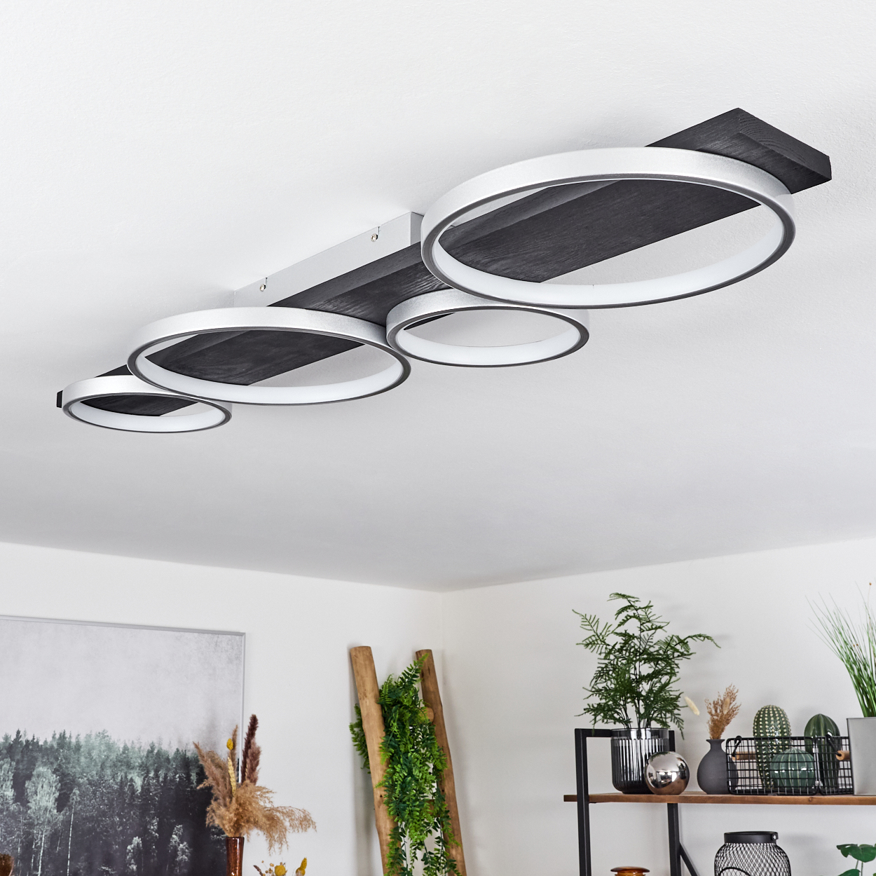 Eine moderne Deckenleuchte mit vier kreisförmigen Lichtern. Darunter ist ein Raum mit Pflanzen, Dekoration, einer Leiter und einem Gemälde.