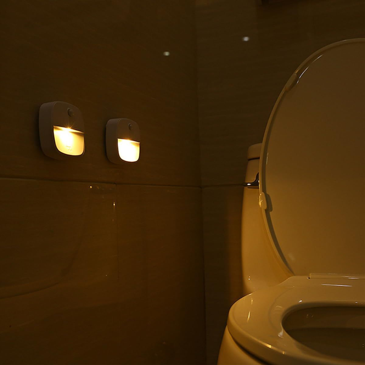 Zwei Wandleuchten beleuchten ein Badezimmer mit einer Toilette. Der Toilettensitz ist hochgeklappt. Die Szene ist schwach beleuchtet.