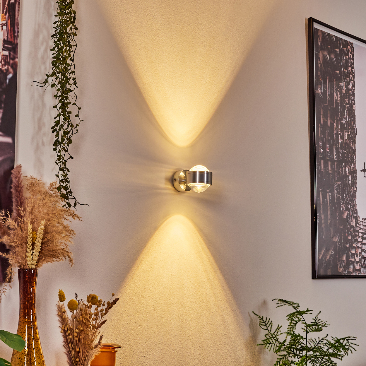 Eine Wandleuchte strahlt Licht in zwei Richtungen aus. Dekorative Pflanzen und ein Bild umgeben die Lampe. Die Wand ist beige.