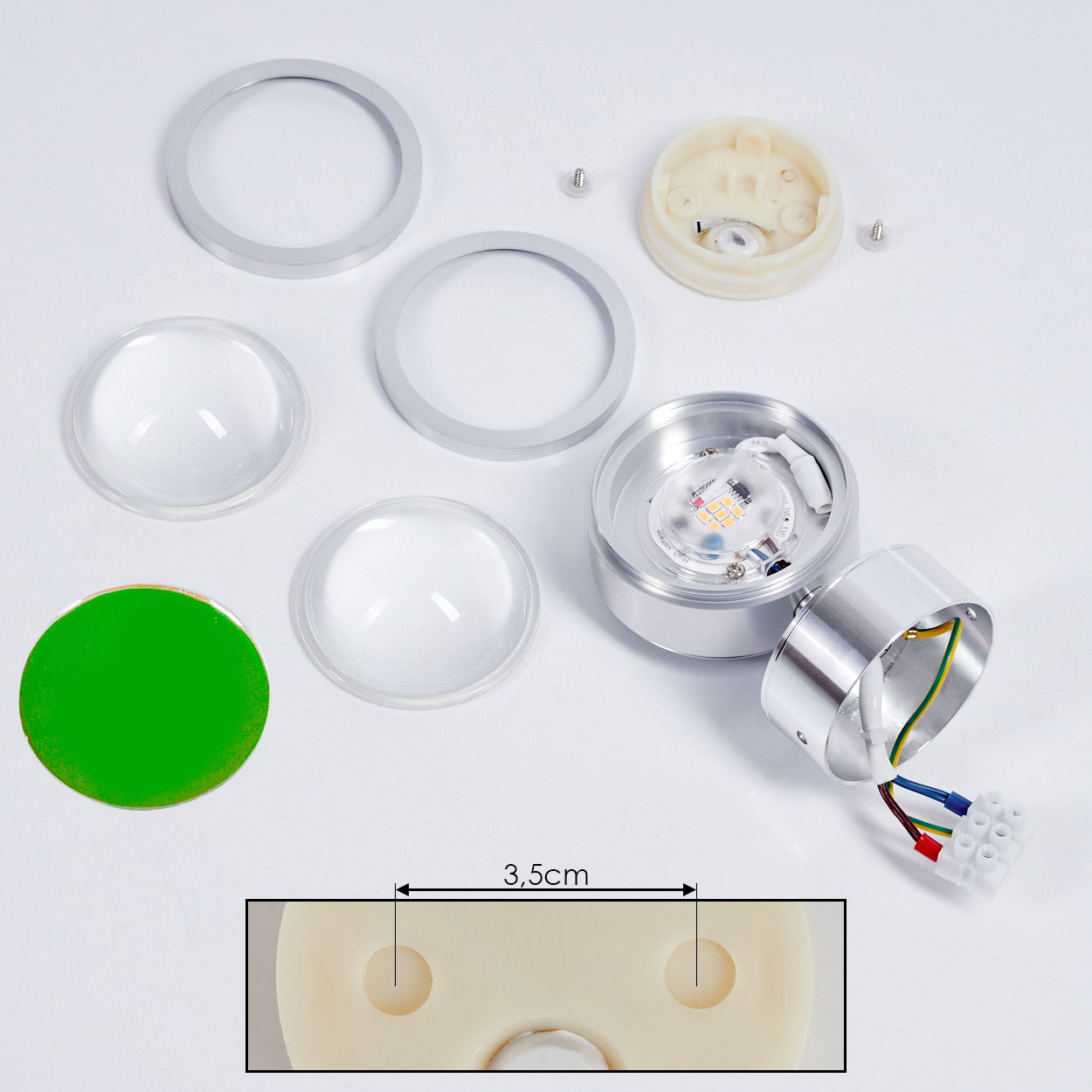 Zerlegte LED-Leuchtkomponenten auf weißer Oberfläche. Enthält Metallringe, Linsen, einen grünen Aufkleber und Kabel.