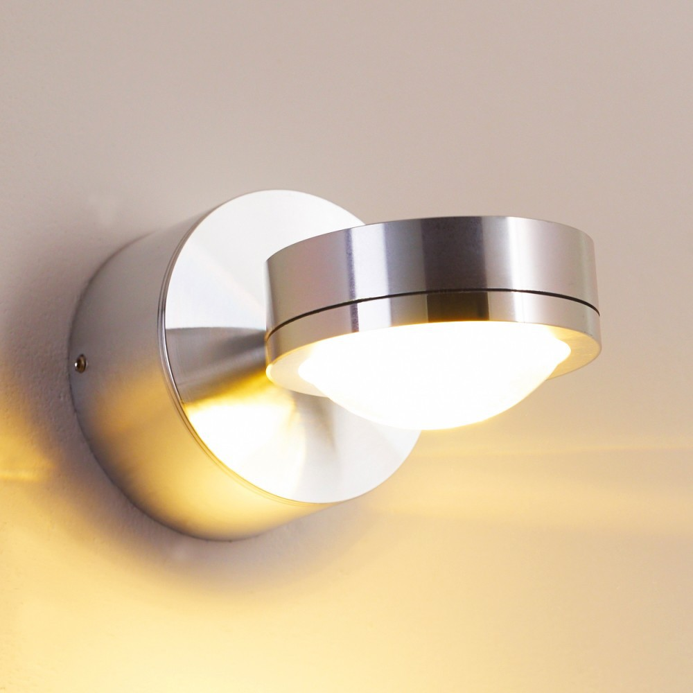 Eine moderne silberne Wandleuchte ist an einer beigefarbenen Wand befestigt und strahlt helles weißes Licht aus. Die Lampe ist kreisförmig mit einem gebogenen Schirm.