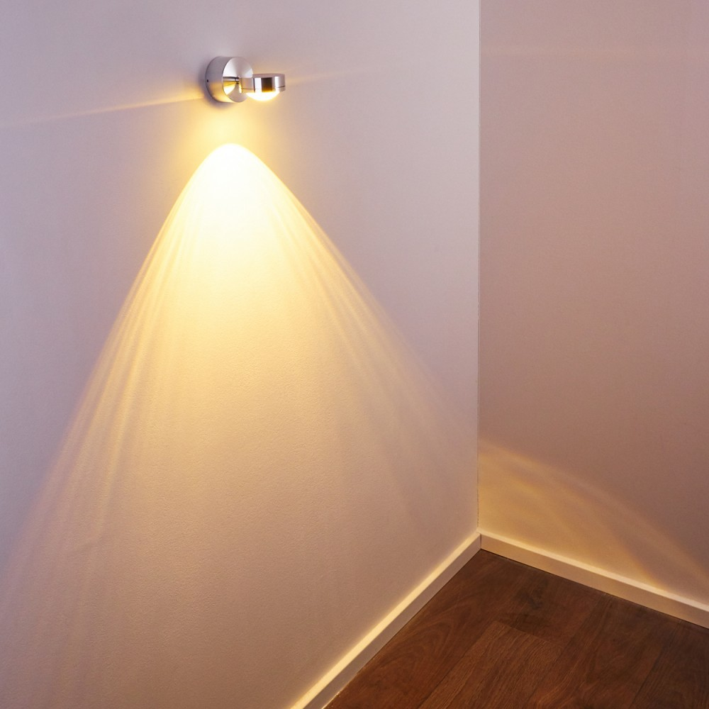 Ein an der Wand montierter Spot beleuchtet eine Wand und einen Holzboden. Das Licht erzeugt eine Kegelform. Die Ecke ist sichtbar.
