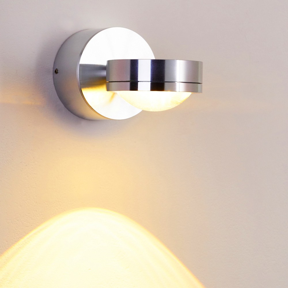 Eine moderne silberne Wandlampe ist an einer hellbeigen Wand befestigt. Die Lampe strahlt ein warmes, helles Licht nach unten aus.