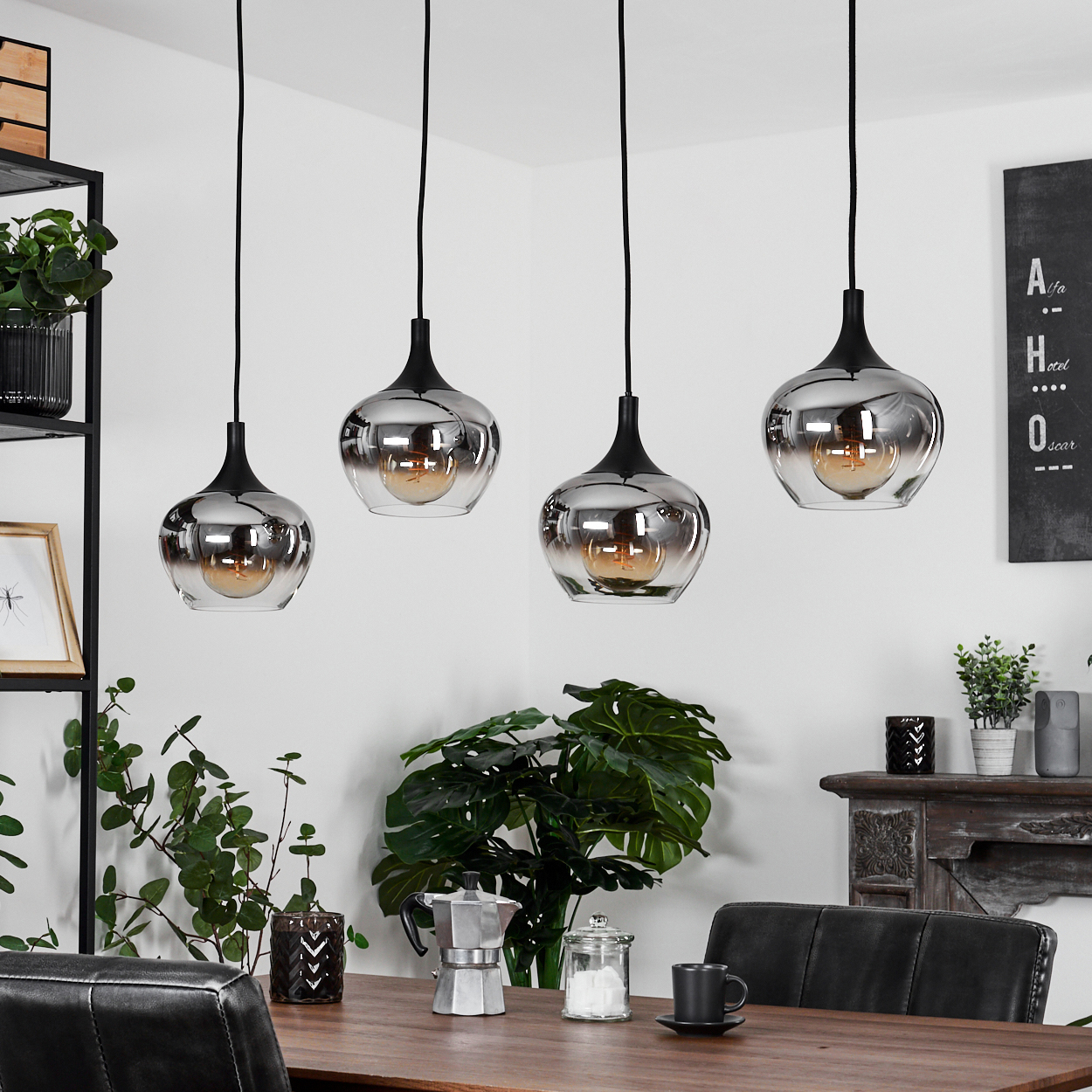 Vier Chrom-Pendelleuchten hängen über einem Holztisch. Pflanzen, eine Kaffeemaschine und ein schwarzer Stuhl sind im Bild.