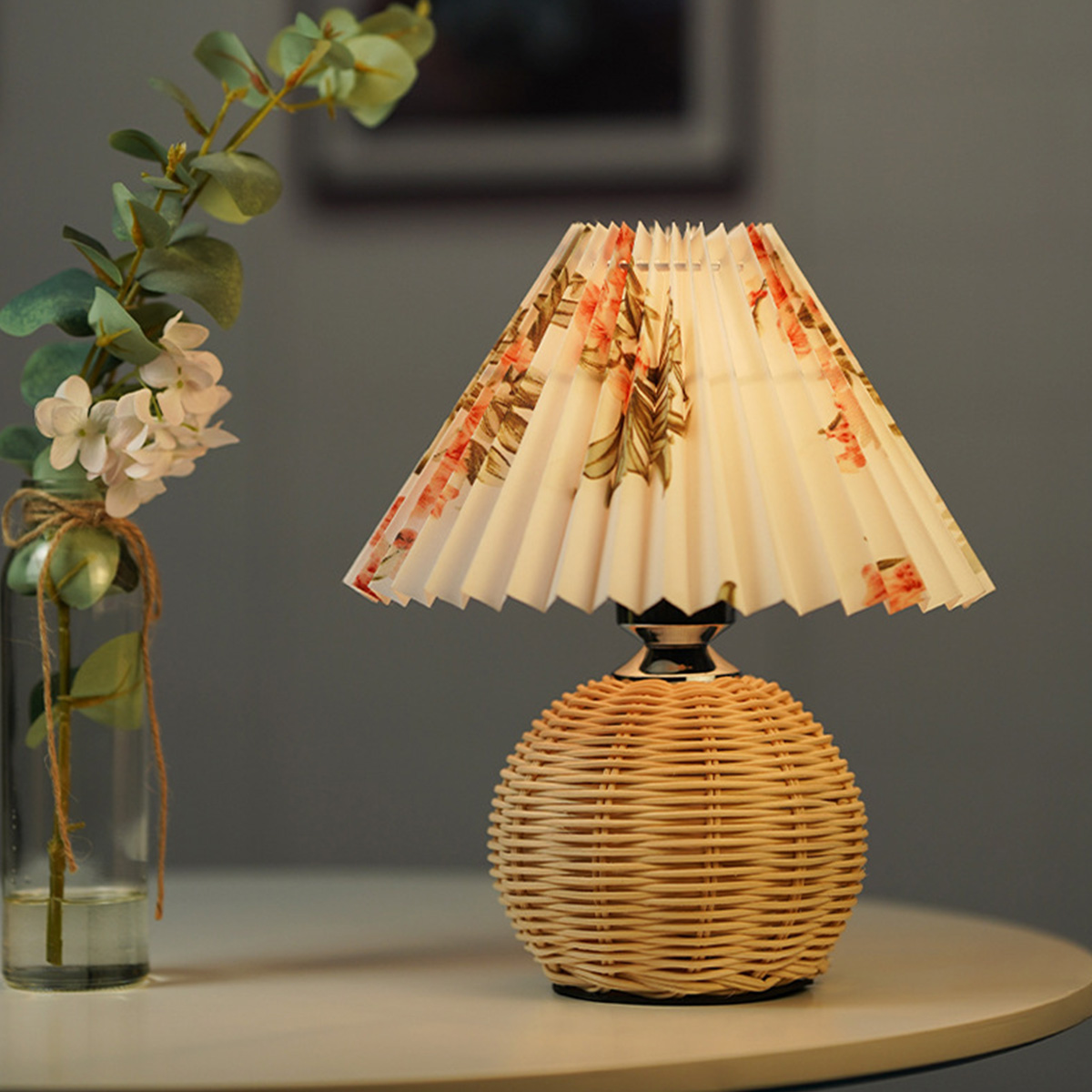 Eine Tischlampe mit Korbfuß und floralem Schirm steht auf einem weißen Tisch. Eine Blumenvase befindet sich links.