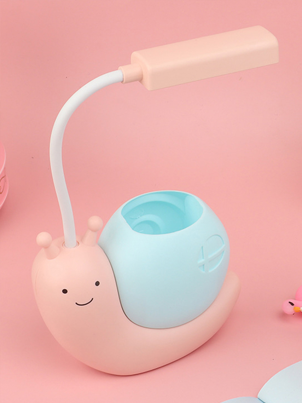 Eine rosa, schneckenförmige Schreibtischlampe mit einem blauen Stifthalter. Die Lampe hat einen weißen, flexiblen Hals und einen hellrosa rechteckigen Kopf.