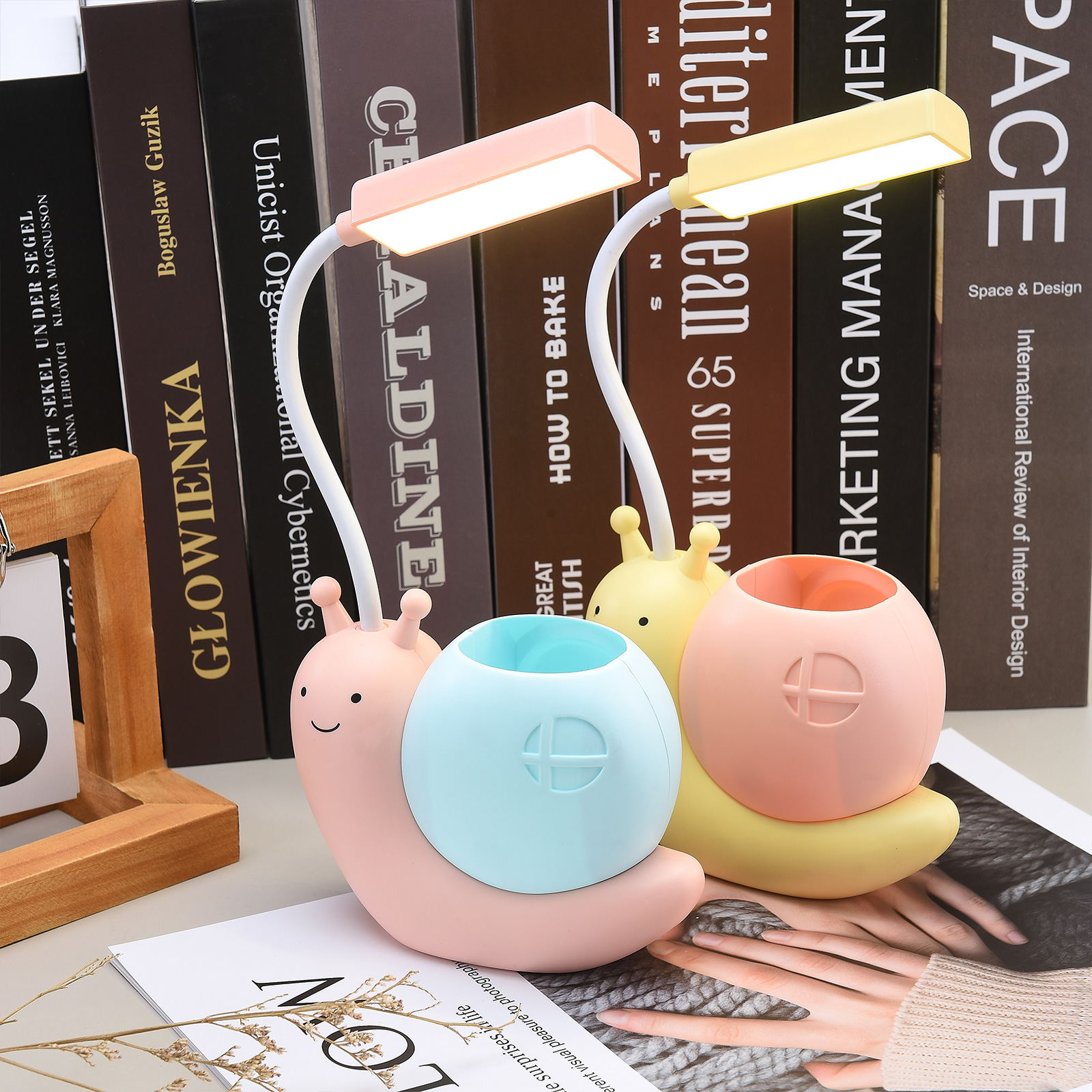 Zwei schneckenförmige Schreibtischlampen, rosa und gelb, mit Stifthaltern. Bücher im Hintergrund und eine Zeitschrift.