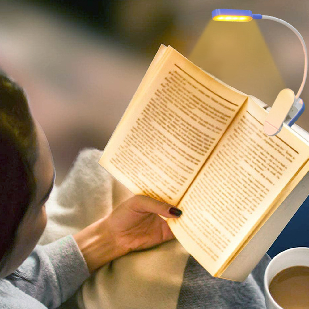 Eine Person liest ein Buch, das von einer kleinen blauen Lampe beleuchtet wird, die am Buch befestigt ist. Eine Tasse Kaffee ist sichtbar.
