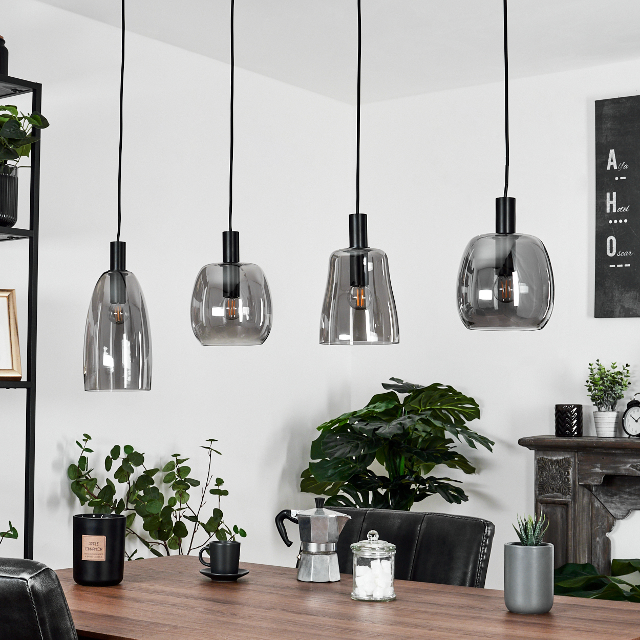 Vier graue Glas-Pendelleuchten über einem Holztisch. Pflanzen, Kaffeemaschine, Kerze und Dekoration sind ebenfalls im Raum vorhanden.