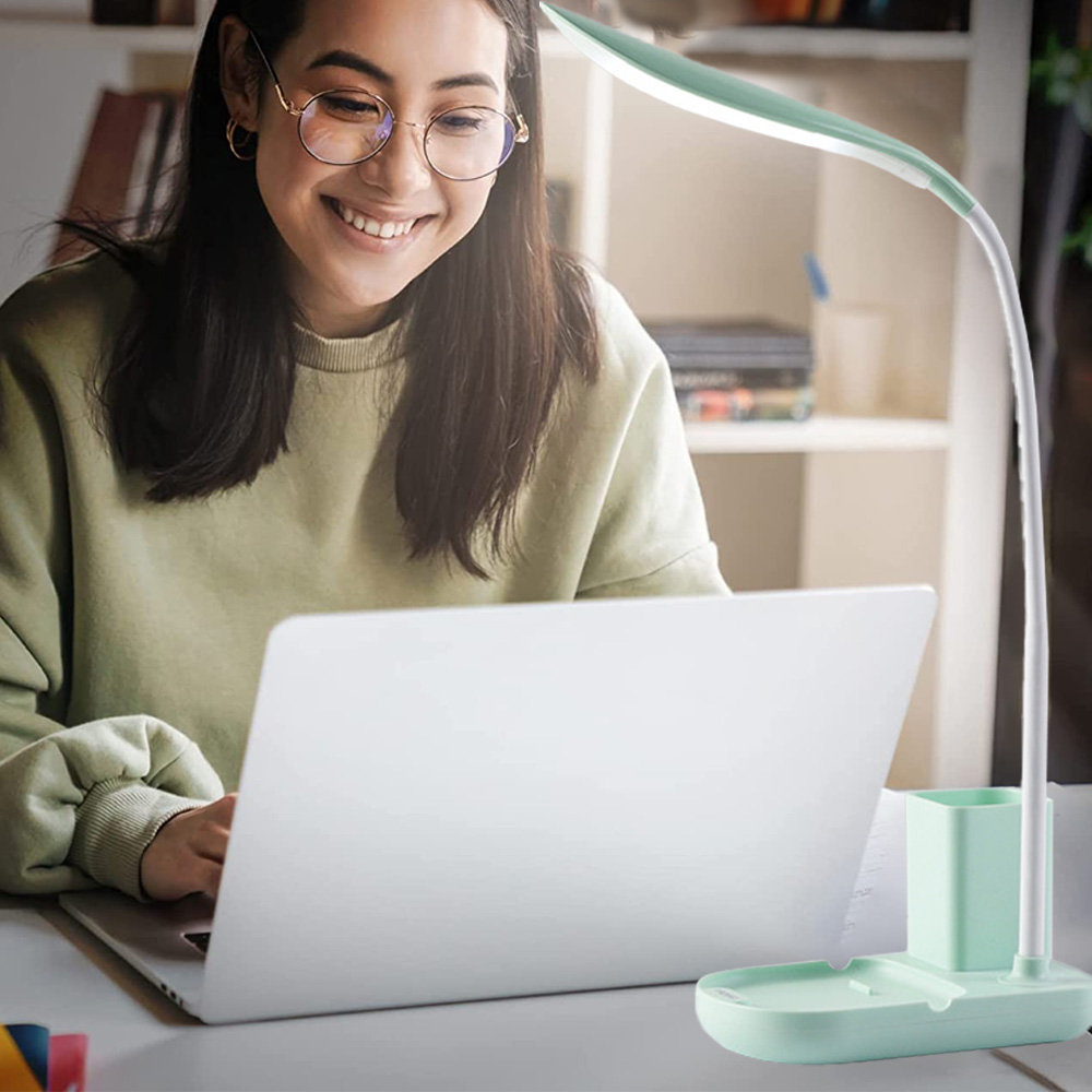 Eine Frau lächelt, während sie einen Laptop benutzt. Rechts ist eine grüne Schreibtischlampe. Im Hintergrund sind Bücherregale zu sehen.