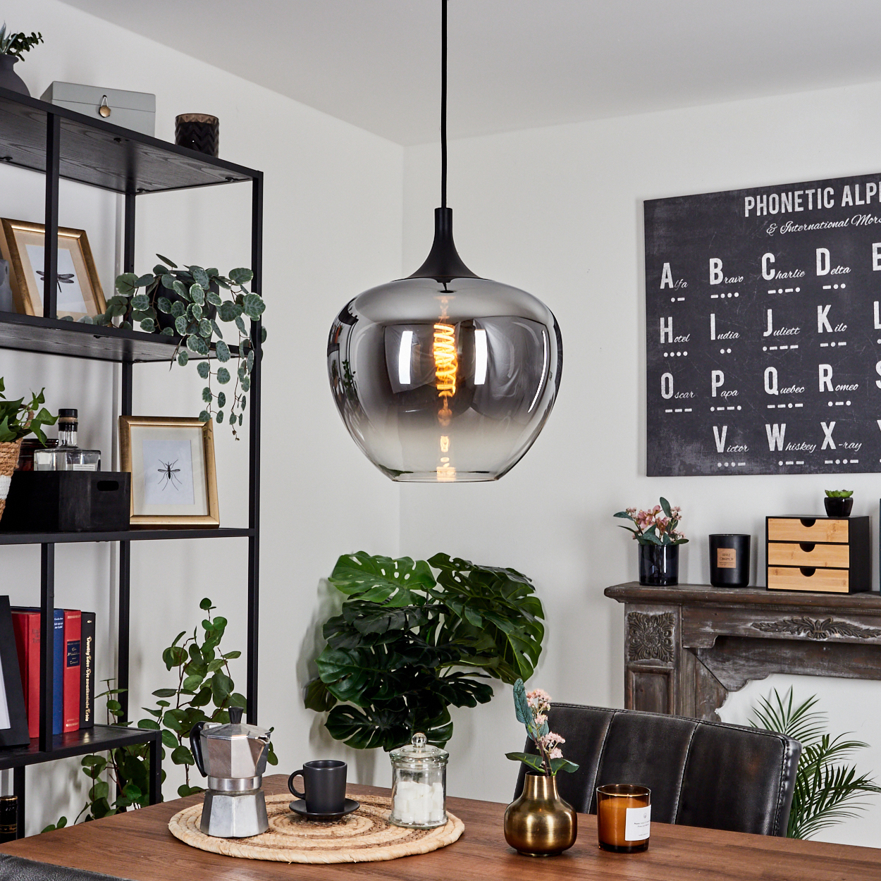 Ein modernes Esszimmer mit großer Pendelleuchte. Tisch mit Kaffeeset, Pflanze und Tafel mit phonetischem Alphabet.