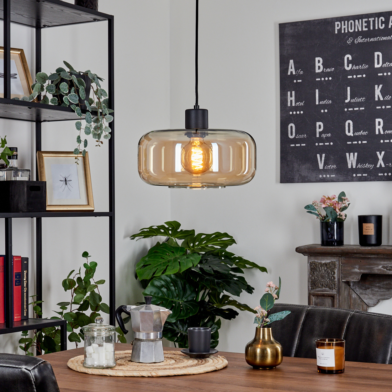 Eine leuchtende, bernsteinfarbene Glas-Hängelampe hängt über einem Holztisch. Eine Kaffeemaschine, Pflanzen und eine Tafel sind im Hintergrund.